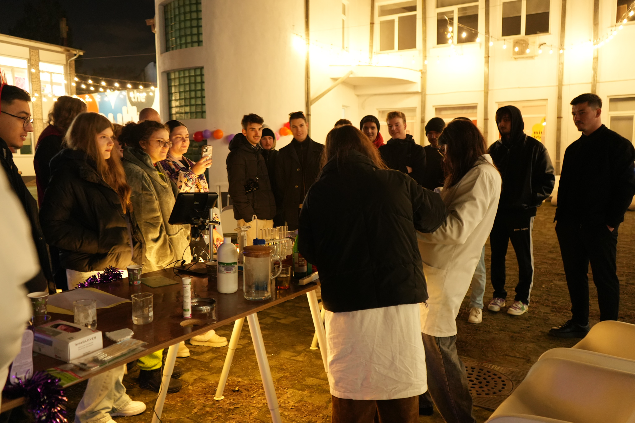 Students and visitors gathered around a demonstration table outdoors at night at the Alacrity Science Fair