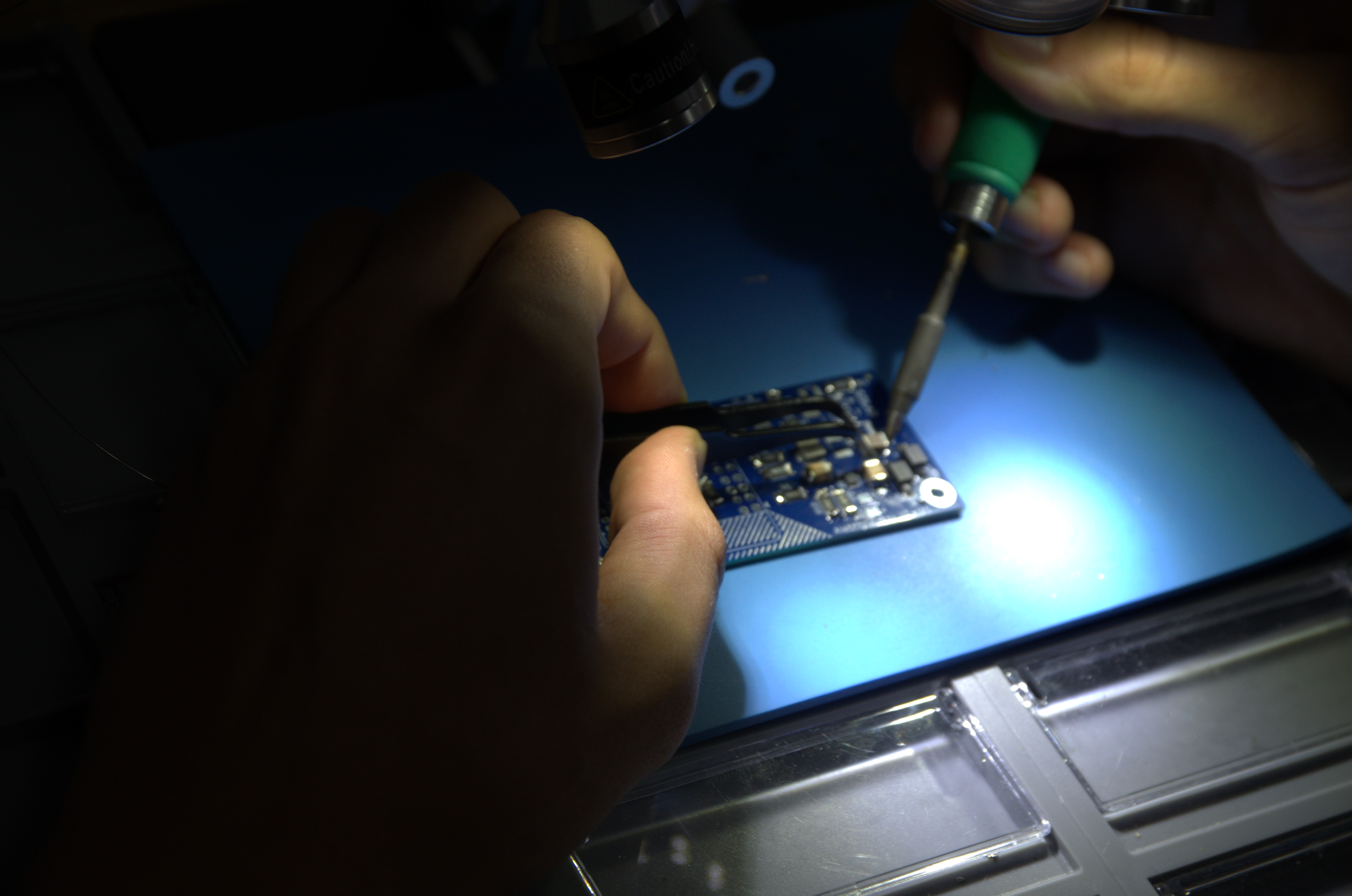 Student soldering a small PCB with tweezers under a lamp