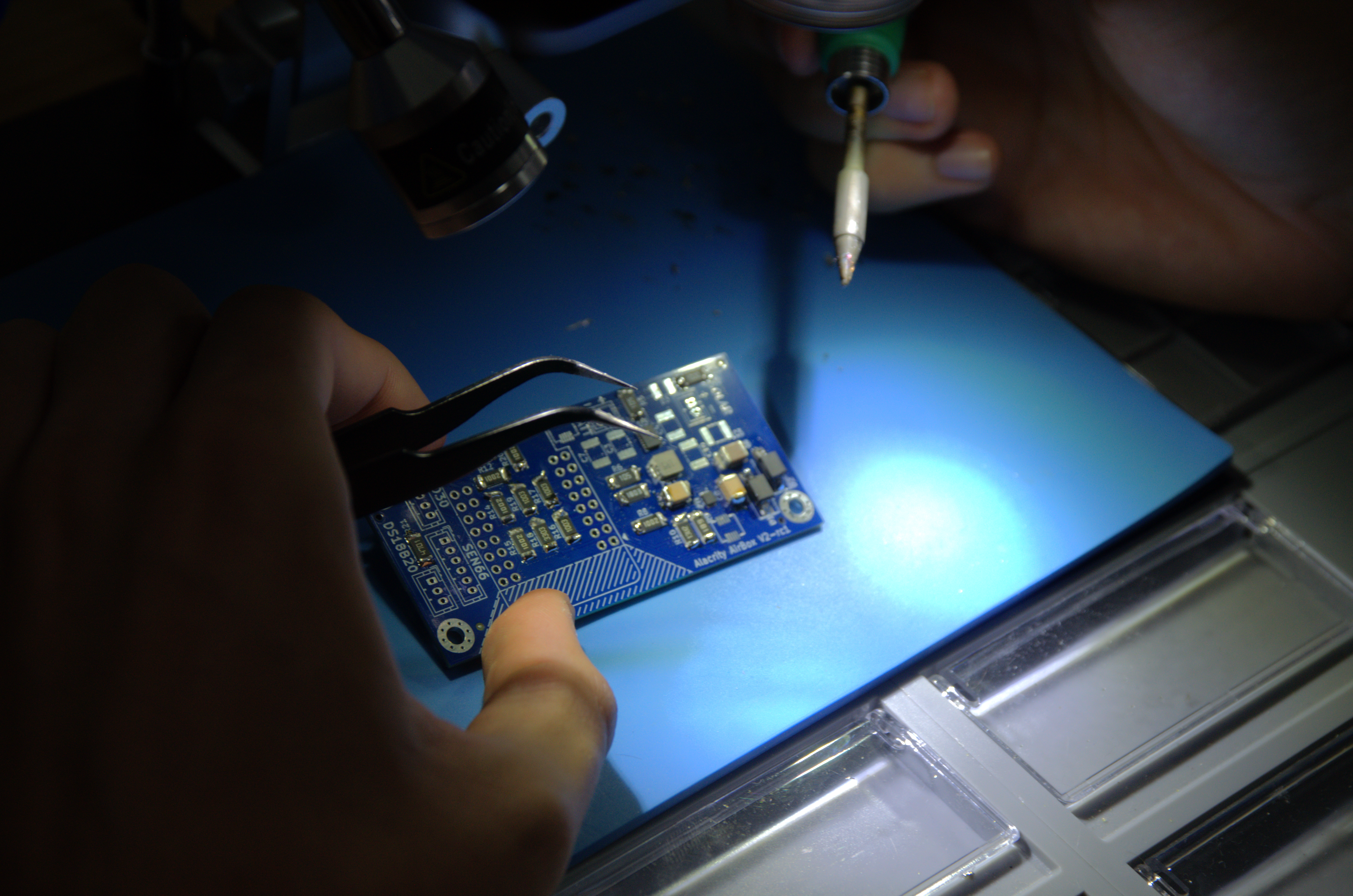 Hands holding a blue PCB with tweezers while soldering under a bright light