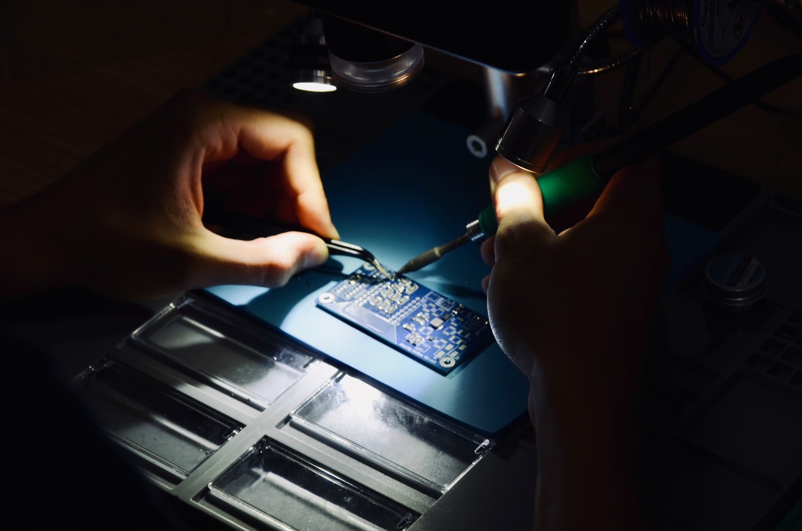 Hands soldering a PCB under a lamp in a dark workshop